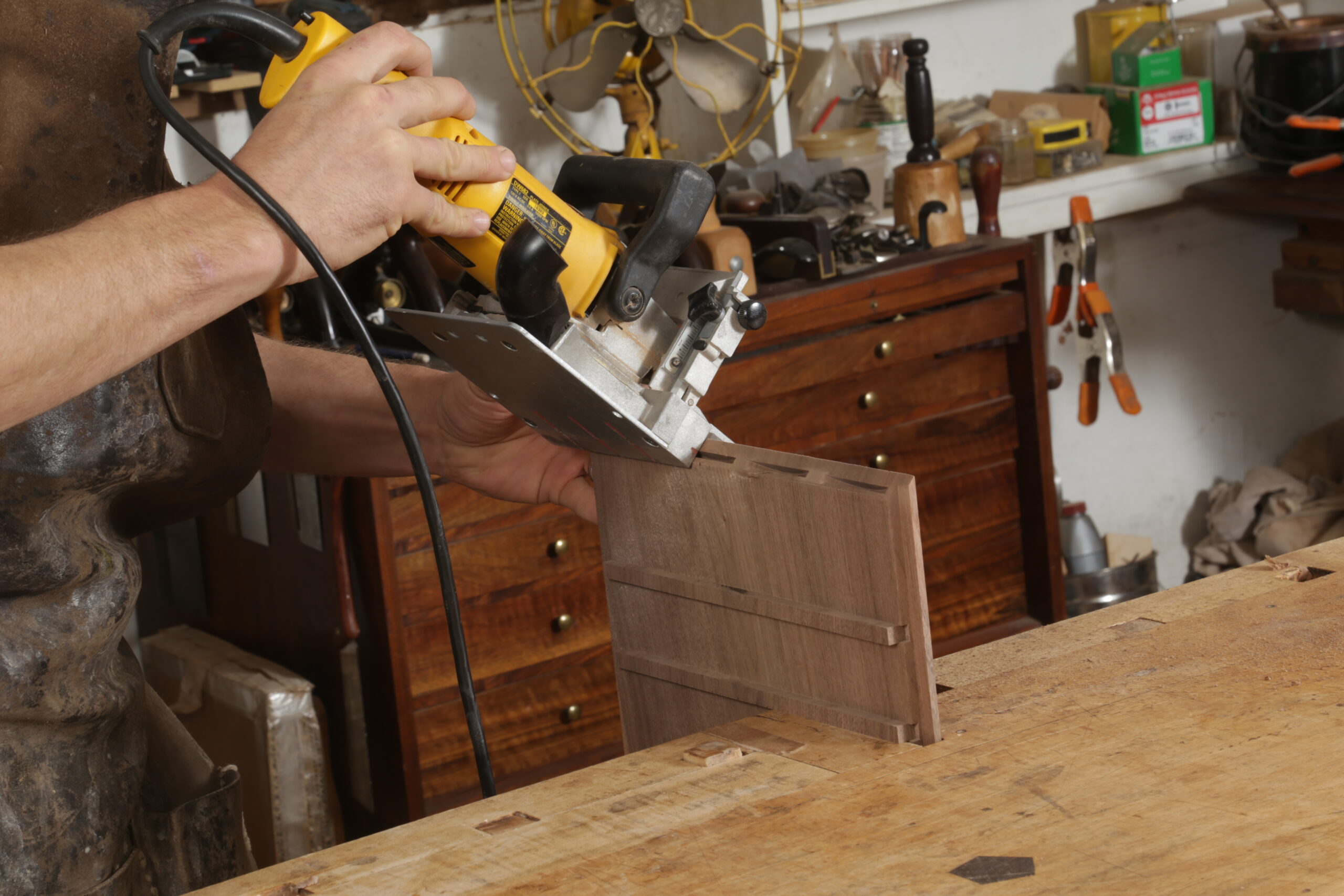 With the box held upright at the workbench, the author is using a biscuit joiner to cut slots in a mitered end. The slots are thin, and there are four of them along the miter. The biscuit joiner is predominantly yellow.