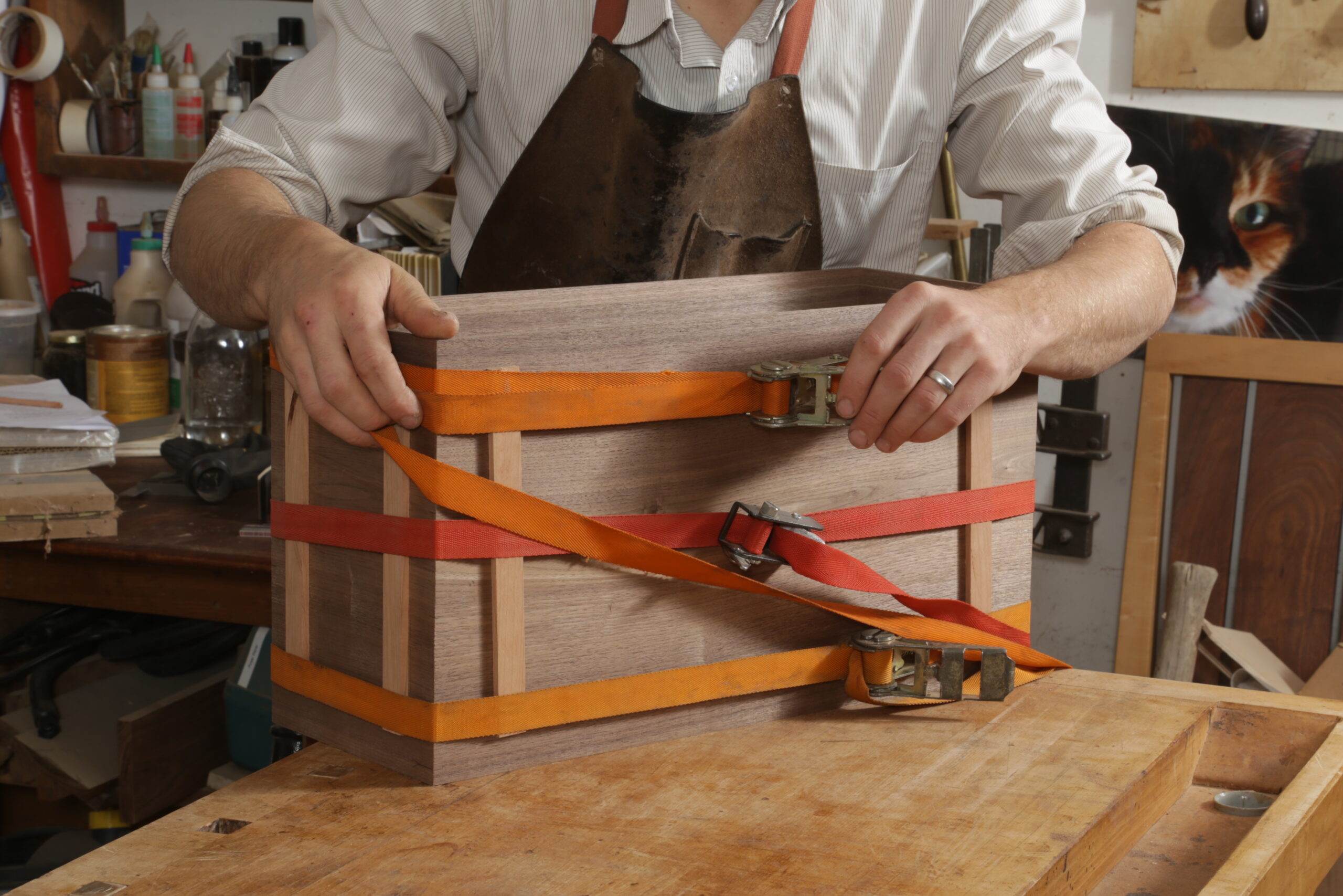 The box carcass is assembled, and the author is clamping it up. He is using two orange ratchet clamps and one red one to apply clamping pressure. At each of the box's corners are two strips of wood between the box and the straps. 